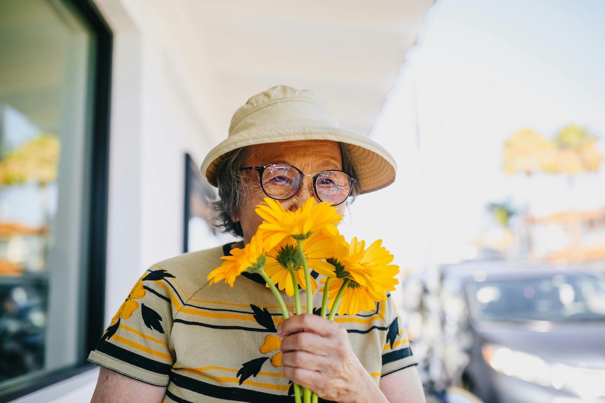 Older woman wearing a bucket hat and glasses holds bright yellow flowers near her face outdoors, smiling softly at the camera.