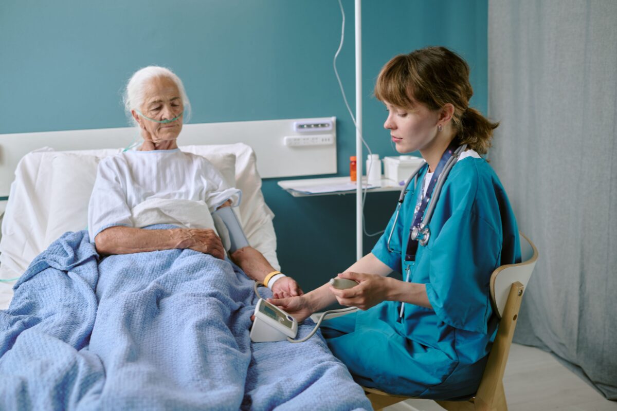 Nurse in teal scrubs checks an elderly patient's blood pressure in a hospital bed with an oxygen tube.