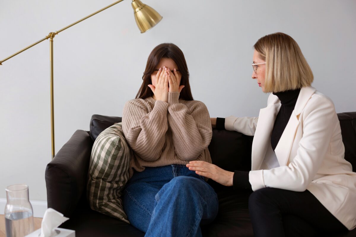 A young woman sits on a black sofa, covering her face with her hands, while a woman in a white blazer consoles her beside her in a living room setting with a brass floor lamp nearby.