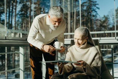 Older man pours coffee for an elderly woman who is seated, looking at a tablet outdoors with trees in the background.