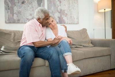 Elderly couple sitting close on a beige sofa, smiling and embracing in a bright living room.