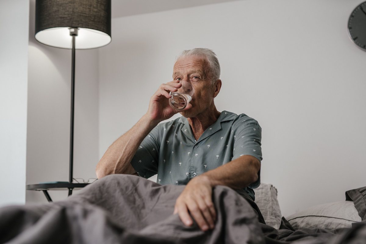 Older man in bed drinking a glass of water at home under a lamp.