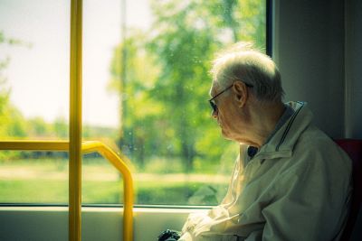 Elderly man with white hair and glasses sits by a window on a bus, looking outside on a sunny day.