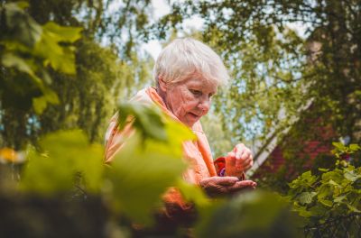 Older woman in an orange jacket tending to plants in a sunlit garden.