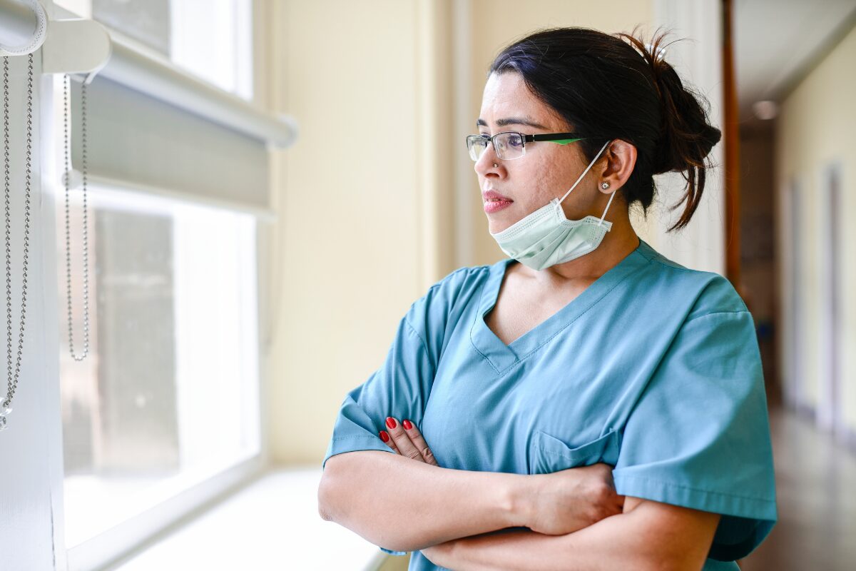Nurse in blue scrubs with a mask under her chin stands with arms crossed, looking out a sunlit window.