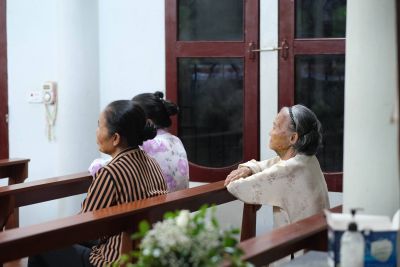 Three elderly women sit on a wooden porch railing, facing left; two wear striped and lavender tops, one in beige, with blurred plants in the foreground.